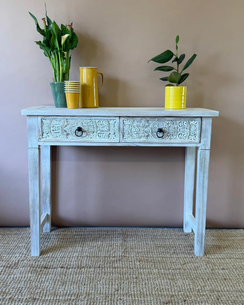 Whitewashed carved console table with two floral-carved drawers and tapered legs, styled with jugs, cups and plants