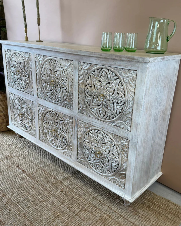 Whitewashed carved sideboard with round floral medallion panels, angled side view, styled with candlesticks and glassware