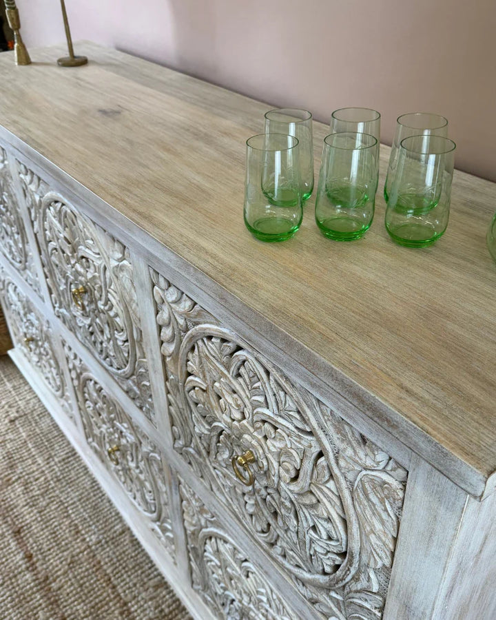 Close-up angled view of a whitewashed carved sideboard with round floral medallion panels, styled with green glassware
