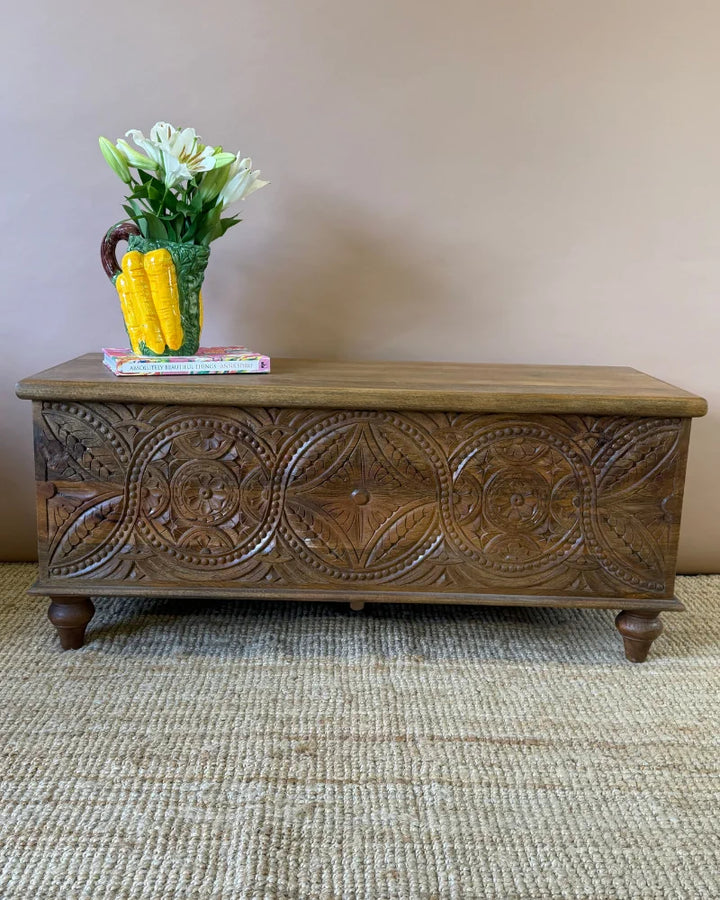 Carved mango wood blanket box with floral medallion carving, turned feet, styled with a ceramic jug of lilies and books on a jute rug