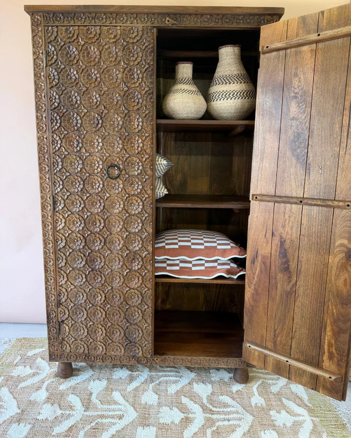 Carved mango wood wardrobe with one door open, revealing shelves styled with woven baskets and patterned cushions