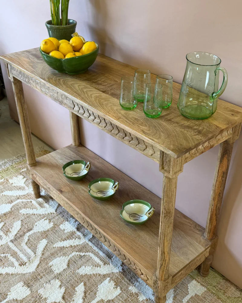 Close-up corner view of a fluted-carved console table styled with a green glass jug and tumblers