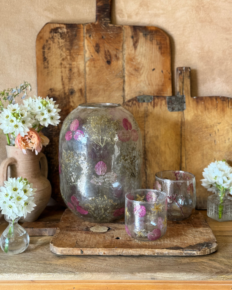 Decorative setting with glass jars, flowers, and a wooden board on a textured wall background.