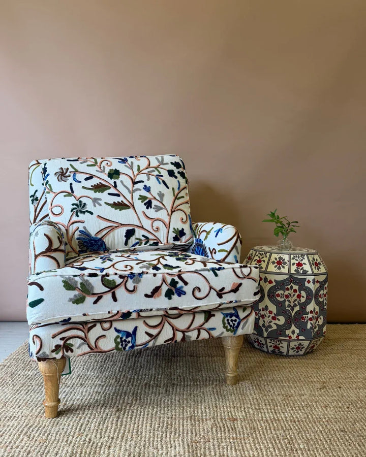 Cream crewel-embroidered armchair positioned beside a hand-painted drum side table, angled view