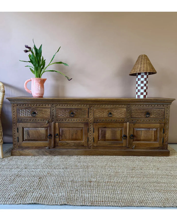 Carved wood sideboard with four drawers over four cupboard doors and leaf and fan carving, styled with a pink jug and checked wicker lamp