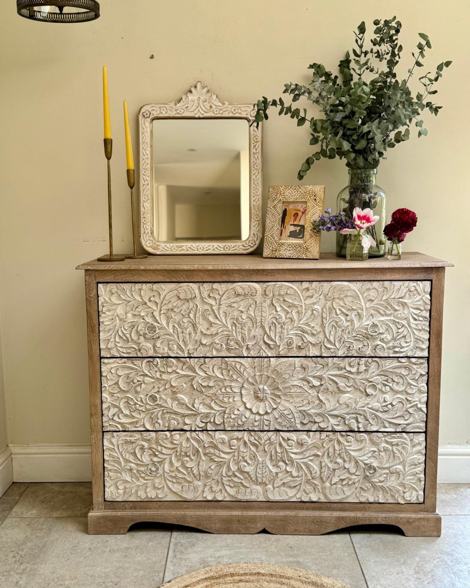 A whitewashed mango wood 3-drawer chest of drawers with hand-carved flower and petal detailing. The chest is accompanied by a mirror, a vase with flowers, and other decorative items.