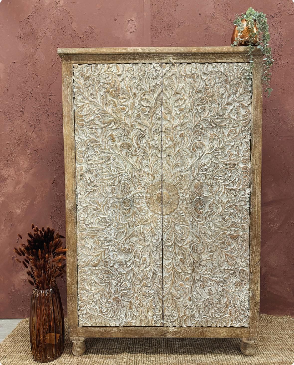 A hand-carved two-door mango wood cabinet with intricate floral designs on the doors, displayed in a room setting with a decorative item on top and a vase with dried plants to the side.