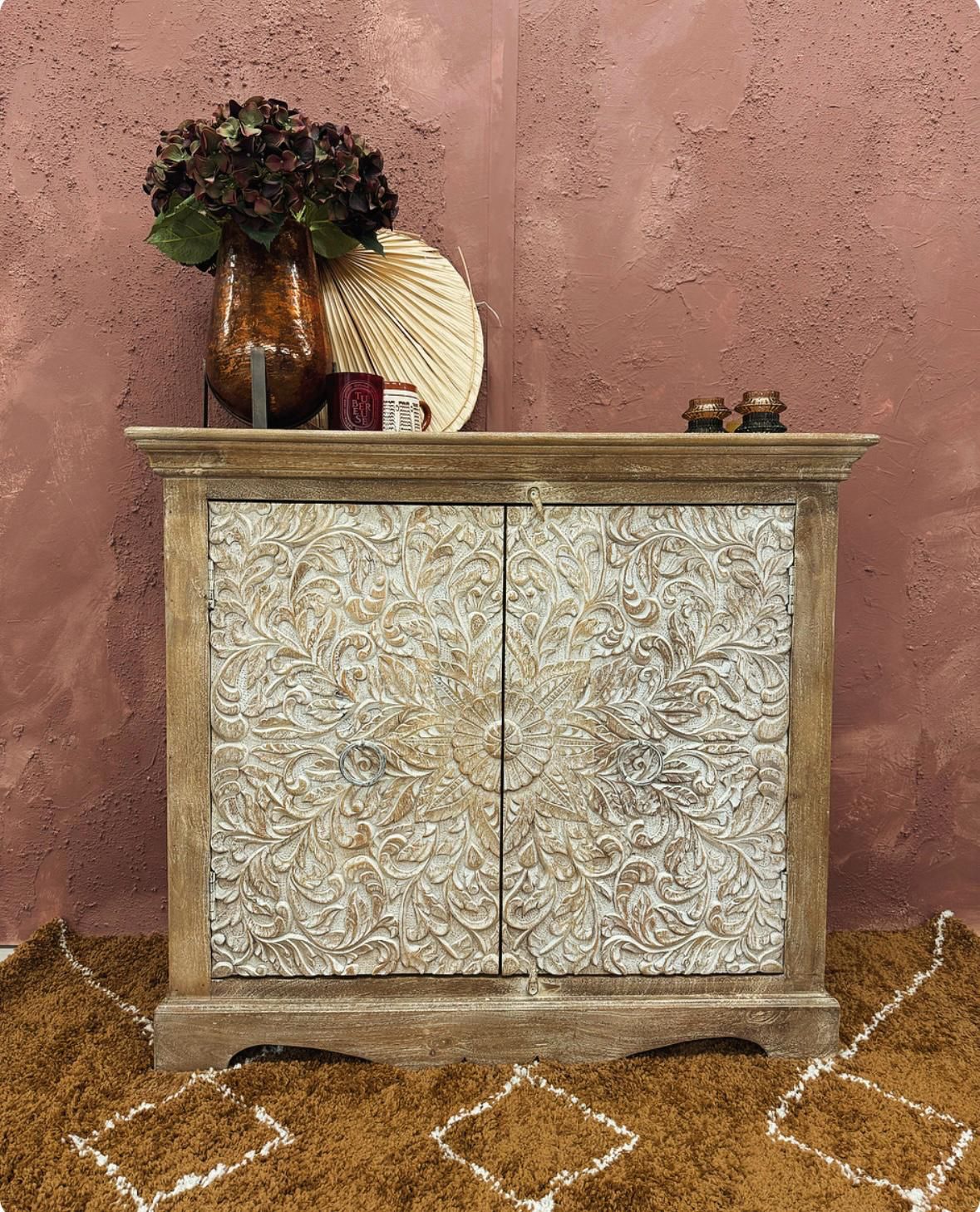 A Two Door Mango Wood Sideboard with hand-carved petal detailing, placed against a textured backdrop, with decorative items on top.