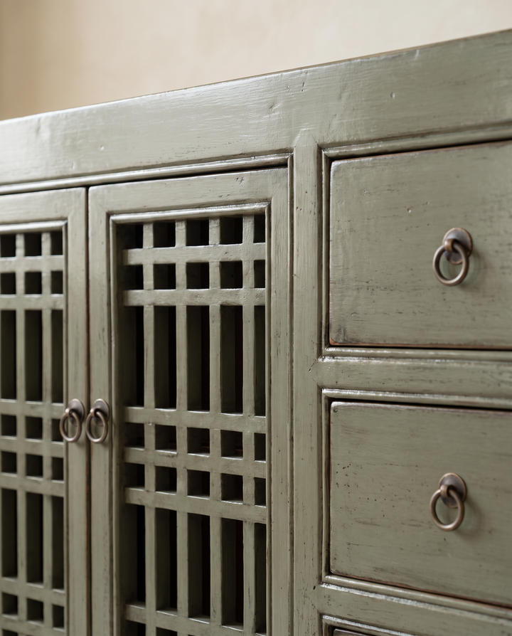 Close-up of a distressed sage green wooden cabinet with lattice-patterned doors and dark metal ring pulls on drawers.