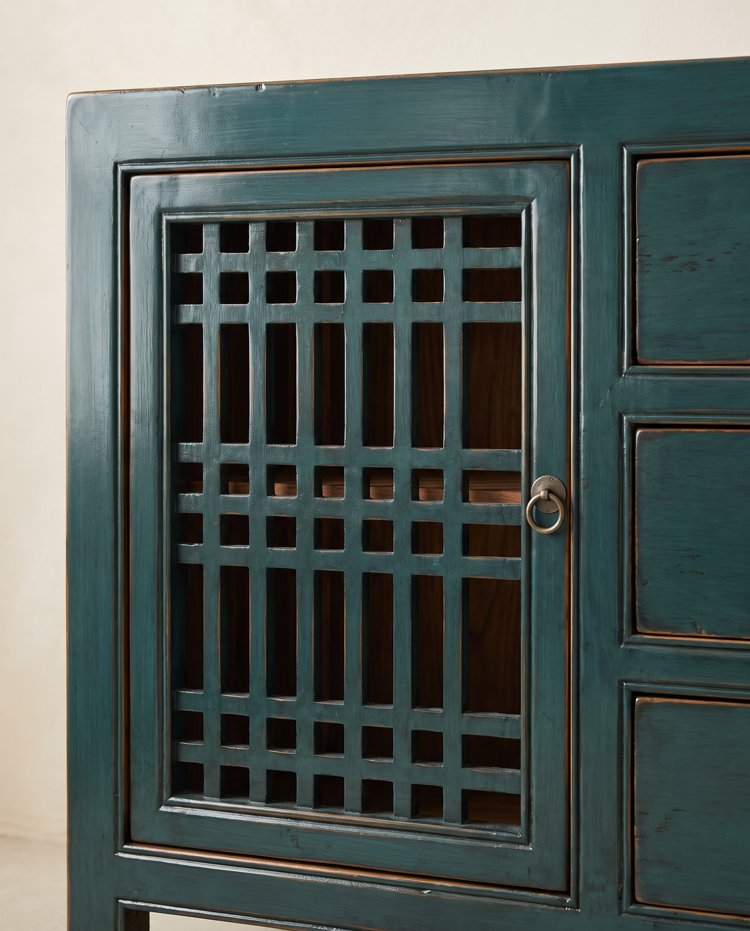 Close-up of a dark teal wooden cabinet with a lattice-patterned door and a round metal ring pull, set against a plain off-white wall.