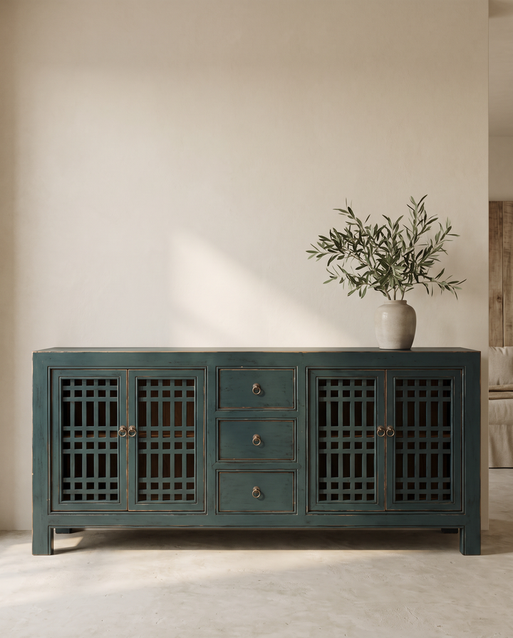 A dark teal-green wooden sideboard cabinet with lattice-patterned doors and three central drawers, styled in a minimalist room with a beige wall and a potted olive branch on top.