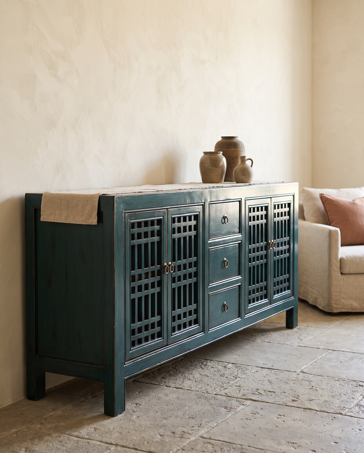 A dark teal wooden sideboard cabinet with lattice-style doors and three drawers, styled in a rustic room next to a beige armchair and vases.