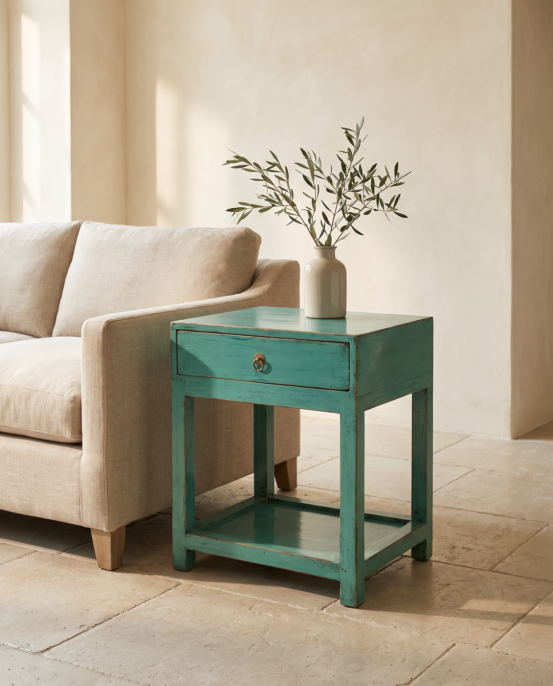 A distressed teal green wooden end table with a single drawer and open lower shelf, styled in a warm neutral room next to a beige sofa with a vase of olive branches on top.