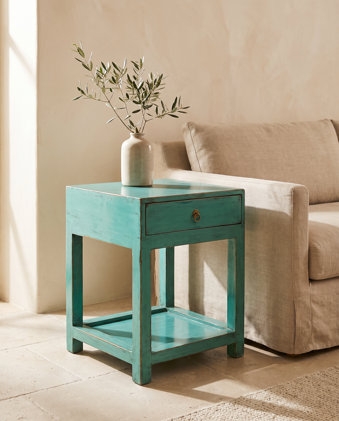 A turquoise painted wooden end table with a single drawer and open lower shelf, styled in a sunlit room next to a beige sofa with a ceramic vase of olive branches on top.