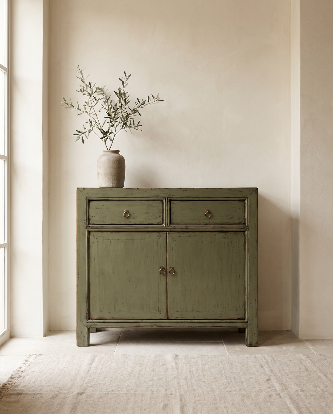 A distressed sage green wooden sideboard cabinet with two drawers on top and two doors below, styled with a ceramic vase of branches in a bright, neutral room.