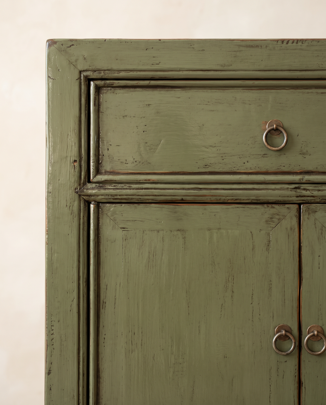 Close-up of a distressed sage green wooden cabinet with a single drawer and two lower doors, featuring round metal ring pulls against a light beige wall.