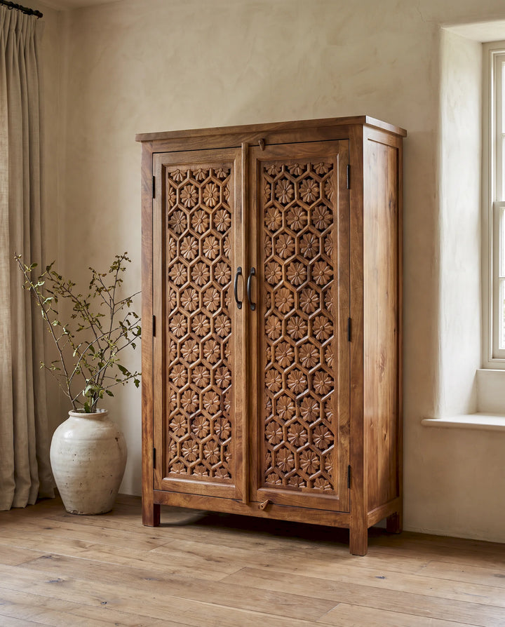 A tall two-door wooden cabinet with intricate carved floral patterns on the doors, shown in a rustic room with beige walls, a window, and a large ceramic vase with dried branches.