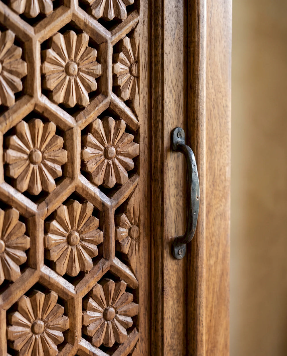 Close-up of a carved wooden cabinet door with a geometric floral pattern and a dark metal handle, set against a softly blurred warm beige background.