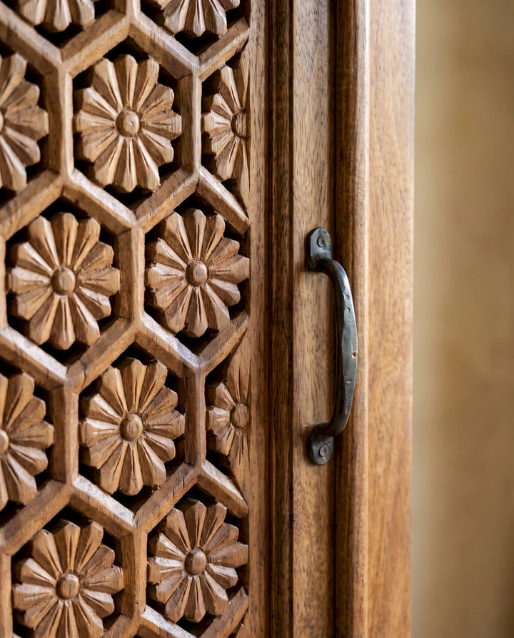 Close-up of a carved wooden cabinet door with a geometric floral pattern and a dark metal handle, set against a softly blurred warm beige background.