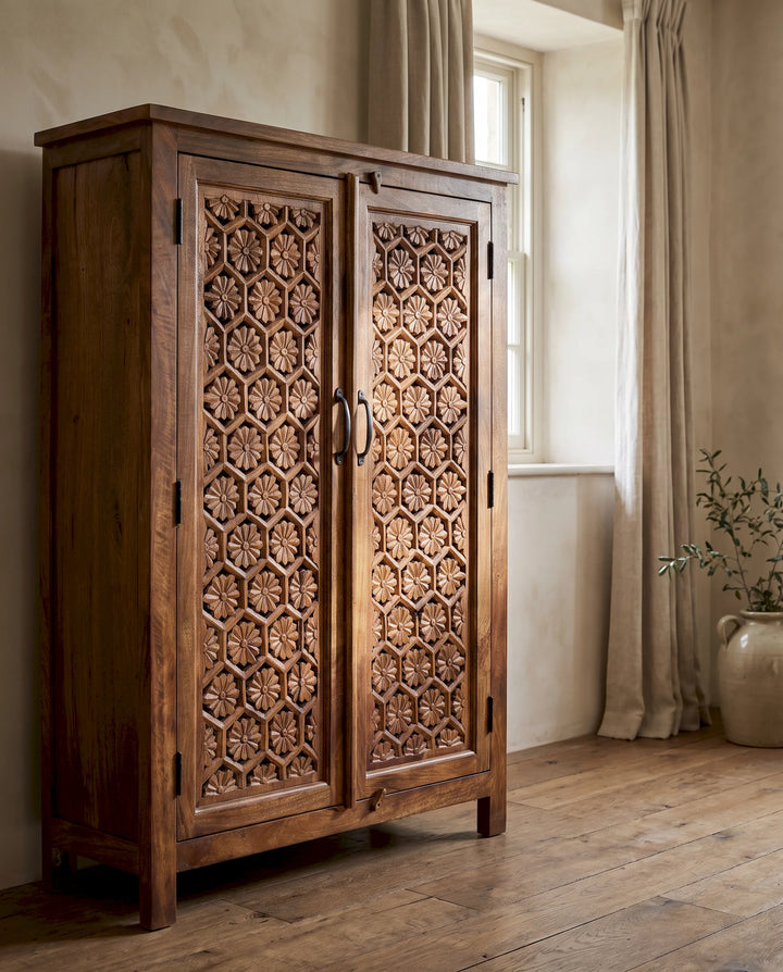 A tall, rustic wooden cabinet with intricate hand-carved floral patterns on its double doors, set in a warm, neutral-toned room with a window and curtains.