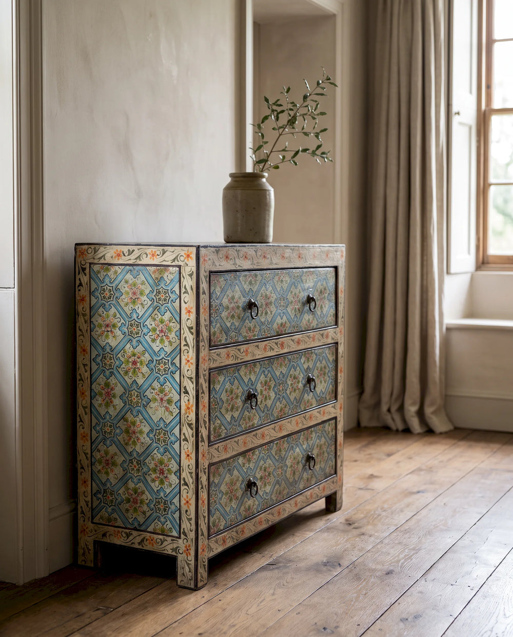 A three-drawer wooden chest of drawers with intricate hand-painted blue, orange, and beige tile patterns, styled in a rustic room next to a window with beige curtains.