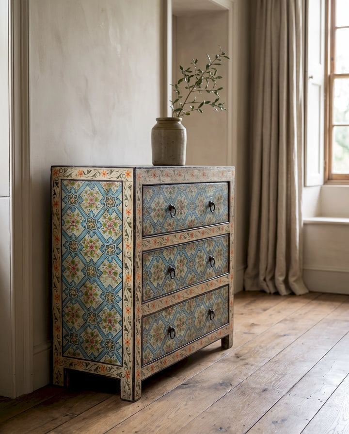 A three-drawer wooden chest of drawers with intricate hand-painted blue, orange, and beige tile patterns, styled in a rustic room next to a window with beige curtains.
