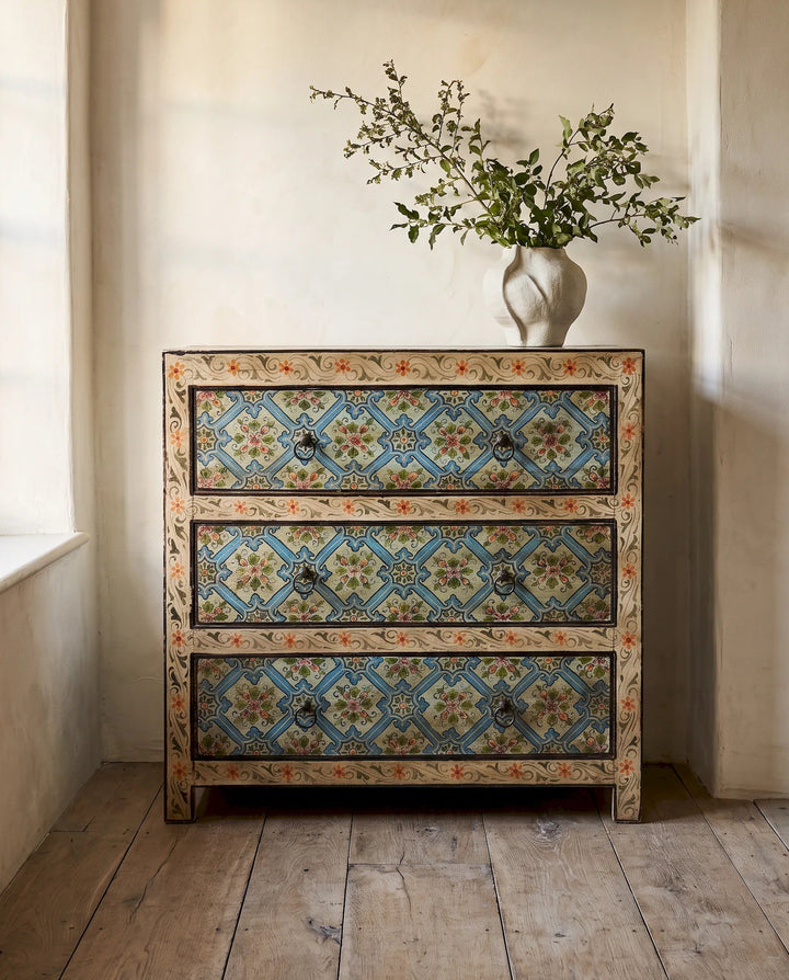 A three-drawer chest of drawers with intricate blue, beige, and orange floral tile patterns, styled in a rustic room with a white vase of greenery on top.